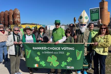 A group of WWURA members all dressed in green hold up a long horizonal banner sign proclaiming Ranked Choice Voting in Ireland since 1921.